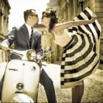 From Italy To Bossa With Love. A well-dressed man kisses a flambouyant women wearing a stunning black and white sun dress hile sitting on a Vespa motorbike on an Italian cobblestone street.
