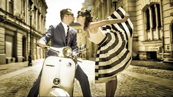 From Italy To Bossa With Love. A well-dressed man kisses a flambouyant women wearing a stunning black and white sun dress hile sitting on a Vespa motorbike on an Italian cobblestone street.