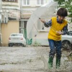 The Waters of March 50th Anniversary. A Young boy plays in a rain puddle while holing a small umbrella.