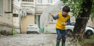 The Waters of March 50th Anniversary. A Young boy plays in a rain puddle while holing a small umbrella.