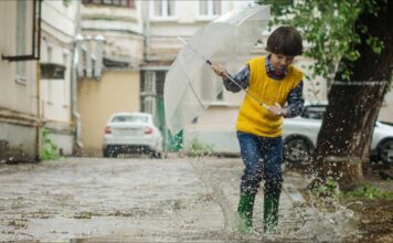 The Waters of March 50th Anniversary. A Young boy plays in a rain puddle while holing a small umbrella.